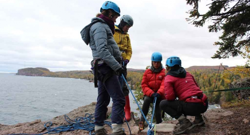 Four people wearing safety gear stand on a cliff above a body of water. Two people are standing, and two are kneeling. 
